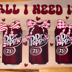 Może przedstawiać: Trzy puszki napoju Dr Pepper z dekoracyjnymi kokardkami. Puszki są ciemnoczerwone z białym tekstem i numerem 23. Kokardy mają czerwono-białe wzory. Tekst "ALL I NEED IS" znajduje się nad puszkami.