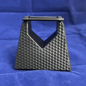 May include: A black plastic stand with a honeycomb pattern and a raised rectangular opening. The stand is designed to hold a small object, such as a phone or a tablet.