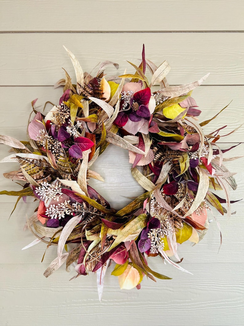 May include: Autumn-themed artificial fall wreath featuring a mix of dried and faux leaves in shades of burgundy, mauve, beige, and gold. The wreath includes various textures and shapes of leaves, along with small artificial flowers and sprigs for added dimension. It's a decorative piece suitable for fall decor, Thanksgiving, or autumnal celebrations.