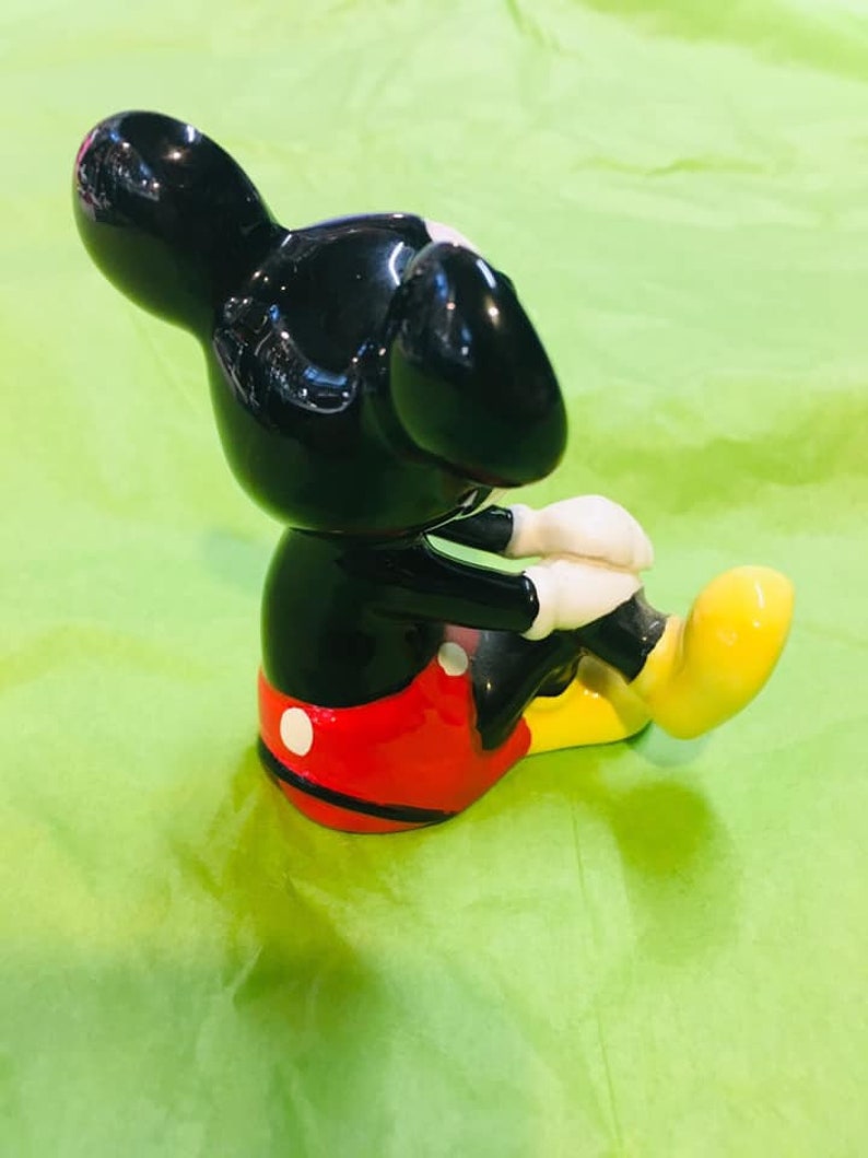 May include: A black ceramic figurine of Mickey Mouse sitting with his legs crossed. He is wearing red shorts with white polka dots and yellow shoes.
