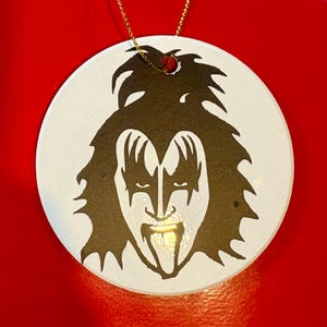 May include: A white, round ornament featuring a black and white illustration of a rock star's face. The image has a gold string for hanging. The rock star has long black hair, eye makeup, and a tongue sticking out.