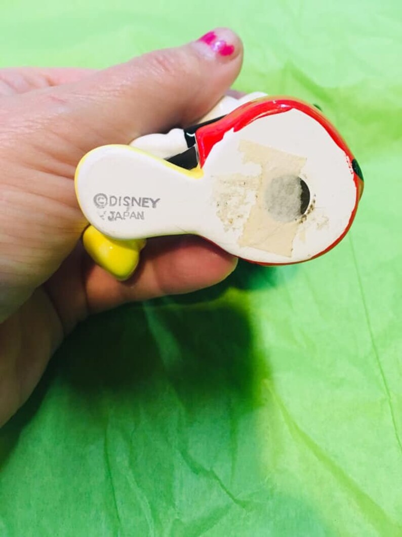 May include: A ceramic figurine of Mickey Mouse wearing a red and yellow outfit. The figurine is made in Japan and has the Disney copyright symbol on the bottom.