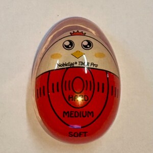 K&ouml;nnte beinhalten: Ein eif&ouml;rmiger K&uuml;chen-Timer mit einer klaren Au&szlig;enh&uuml;lle. Die Innenfl&auml;che zeigt eine Cartoon-H&uuml;hnergrafik mit dem Text NobleEgg TIMER Pro. Darunter befindet sich eine rote kreisf&ouml;rmige Skala mit den Beschriftungen HARD, MEDIUM und SOFT.