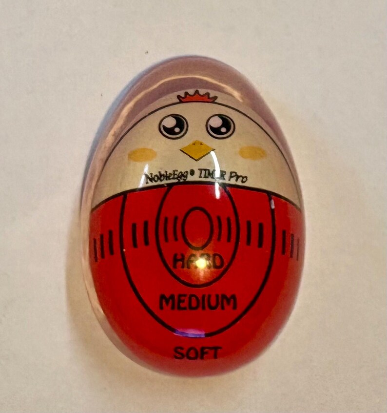 K&ouml;nnte beinhalten: Ein eif&ouml;rmiger K&uuml;chen-Timer mit einer klaren Au&szlig;enh&uuml;lle. Die Innenfl&auml;che zeigt eine Cartoon-H&uuml;hnergrafik mit dem Text NobleEgg TIMER Pro. Darunter befindet sich eine rote kreisf&ouml;rmige Skala mit den Beschriftungen HARD, MEDIUM und SOFT.