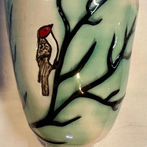 May include: A hand-painted ceramic cup with a bird perched on a branch. The cup features a light green and white background with black branches and a red-headed bird. The cup has a bulbous base and a flared rim.