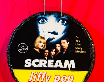 Könnte beinhalten: Ein schwarzer und gelber Jiffy Pop Popcorn-Behälter mit Scream-Filmthema. Der Behälter zeigt eine Nahaufnahme der Ghostface-Maske und den Text "Do You Like Scary Movies?" und "SCREAM".