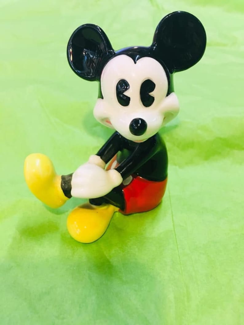 May include: A black and white ceramic figurine of Mickey Mouse sitting with his legs crossed. He is wearing red shorts and yellow shoes.