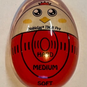 May include: An egg-shaped kitchen timer with a glossy finish featuring a cartoon chicken face at the top. The center displays the text NobleEgg TIMER Pro with red concentric circles and the labels HARD, MEDIUM, and SOFT printed in black text.