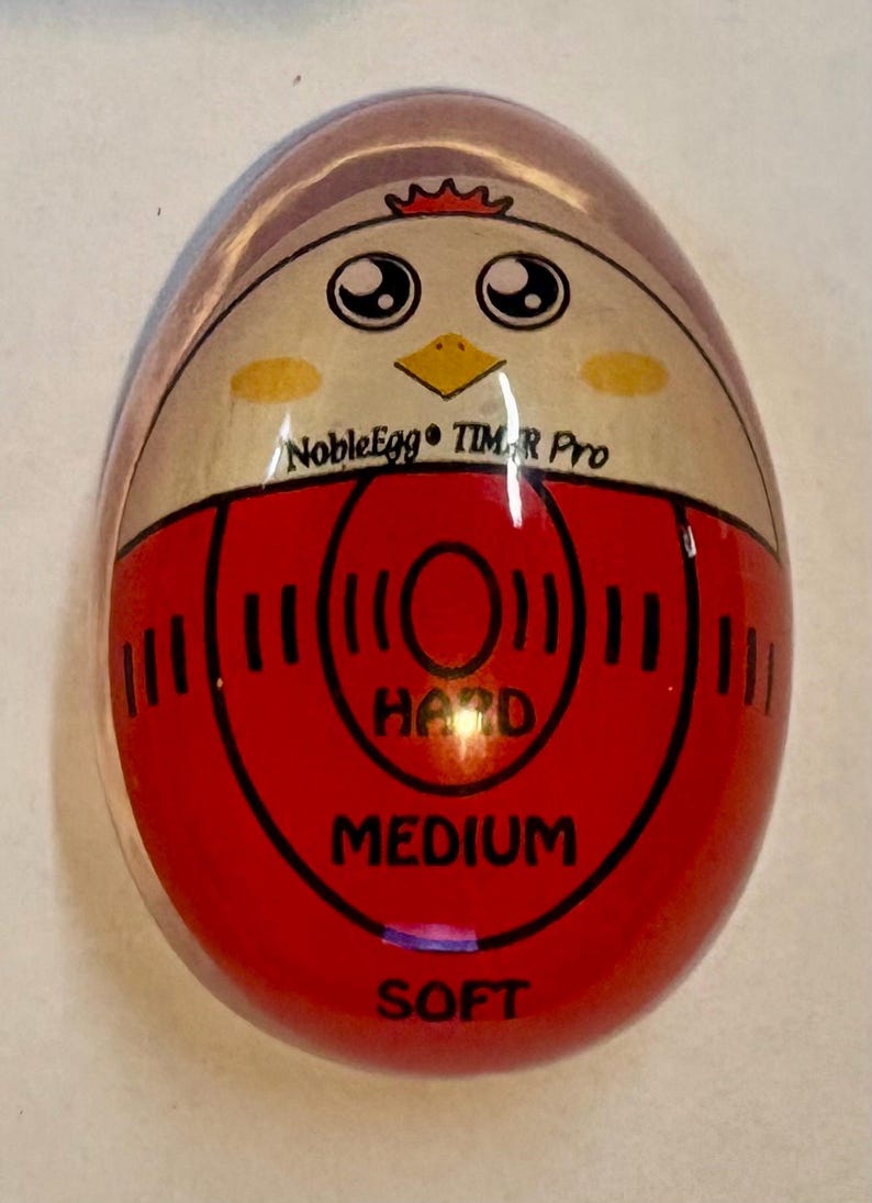 K&ouml;nnte beinhalten: Ein eif&ouml;rmiger K&uuml;chen-Timer mit gl&auml;nzender Oberfl&auml;che und einem Cartoon-H&uuml;hnergesicht an der Oberseite. In der Mitte befindet sich der Schriftzug NobleEgg TIMER Pro mit roten konzentrischen Kreisen sowie den Beschriftungen HARD, MEDIUM und SOFT.