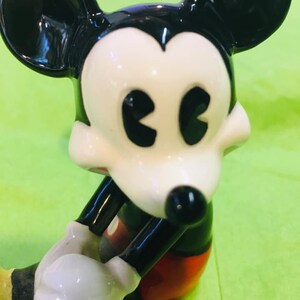 May include: A ceramic figurine of Mickey Mouse in his classic red shorts and yellow shoes. He is sitting with his arms crossed and has a black and white face with large, black eyes.