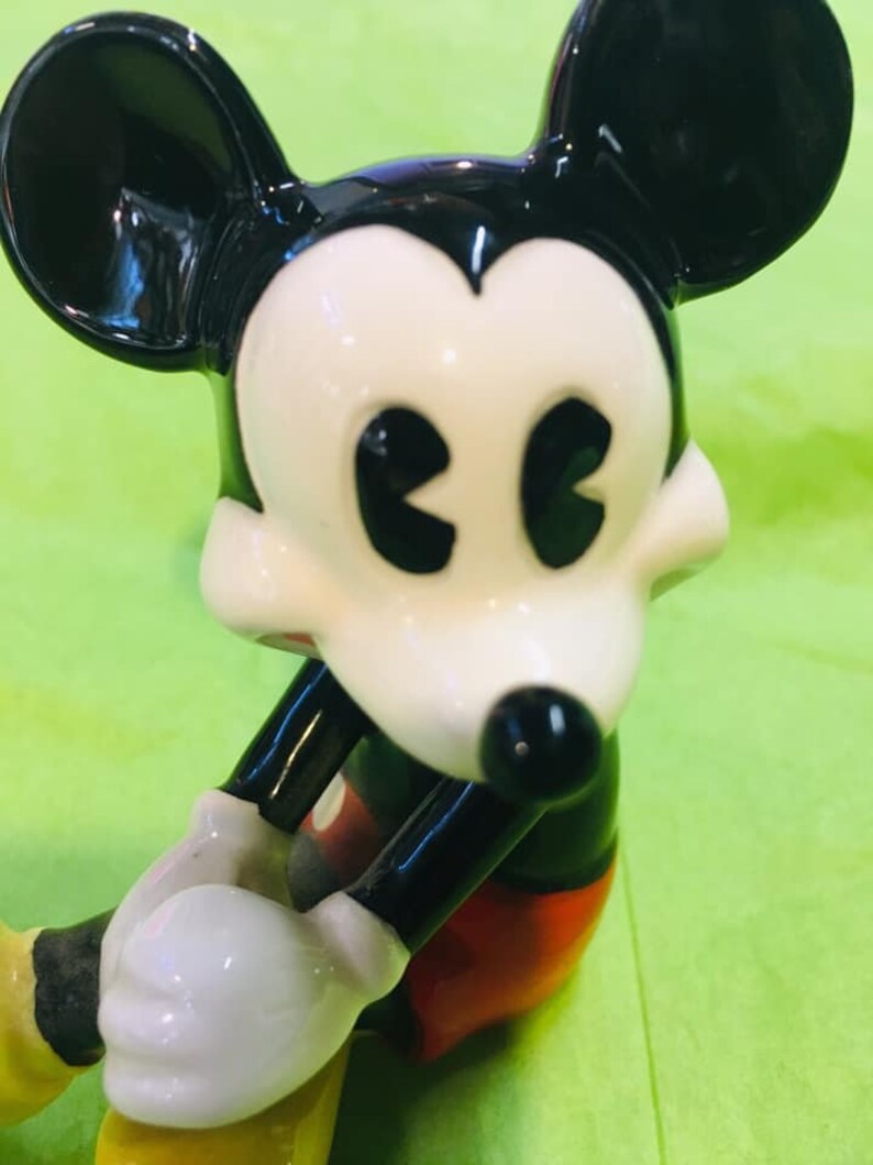 May include: A ceramic figurine of Mickey Mouse in his classic red shorts and yellow shoes. He is sitting with his arms crossed and has a black and white face with large, black eyes.