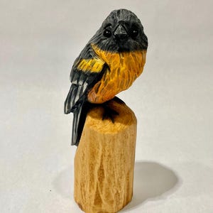 May include: A hand-carved wooden figurine of a bird with black plumage on its head and wings, featuring a bright orange chest and belly. The bird is perched atop a cylindrical, light-colored wooden log base with visible carved textures.