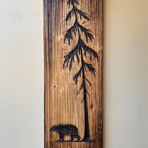 May include: A rectangular wooden wall art piece with a carved design. The artwork features a tall tree and a bear silhouette. The wood has a natural grain and a dark, burnt border, creating a rustic aesthetic.