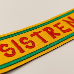 May include: A yellow fabric patch with a green border and red embroidered text that reads "SISTREN".