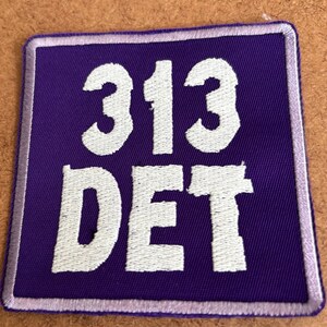 May include: A square purple patch with white embroidered text. The text reads "313 DET" in a bold, sans-serif font. The patch has a thin, light purple border.
