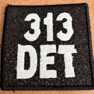 May include: Black square patch with white embroidered text reading "313 DET". The patch has a glittery texture and a black border. The patch is likely an emblem or a badge.