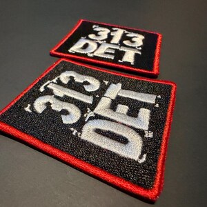May include: Two rectangular embroidered patches featuring the text 313 DET in white thread on a black background. Each patch is bordered by a thick red stitched edge. The patches are positioned at an angle, showing the textured embroidery.