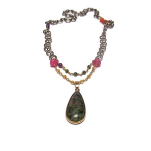 Puede incluir: Un collar de dos hilos con un colgante de piedra preciosa verde en forma de lágrima. El collar presenta cuentas rosas, verdes y doradas.
