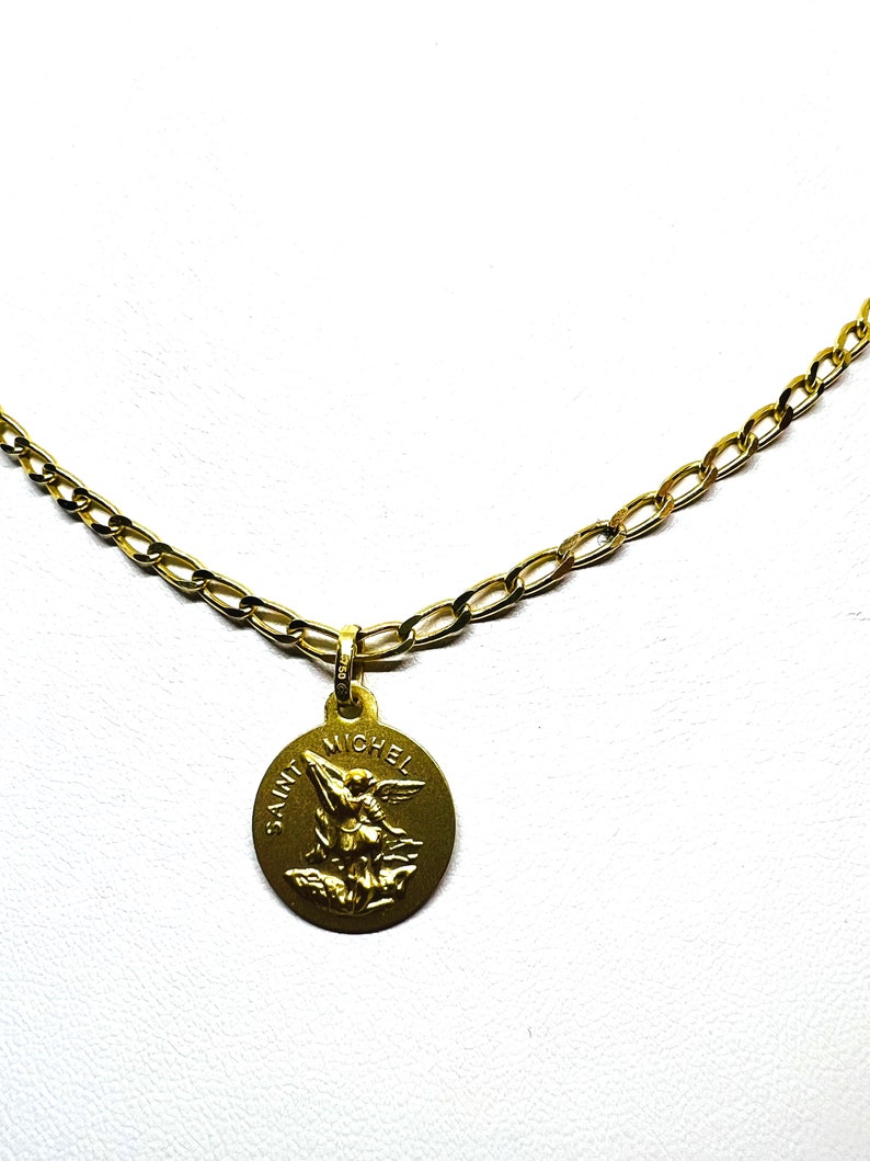 Puede incluir: Un collar dorado con un colgante redondo con la inscripci&oacute;n "SAINT MICHEL" y una imagen de un &aacute;ngel. La cadena es de dise&ntilde;o de eslabones. El colgante tiene un aspecto envejecido.