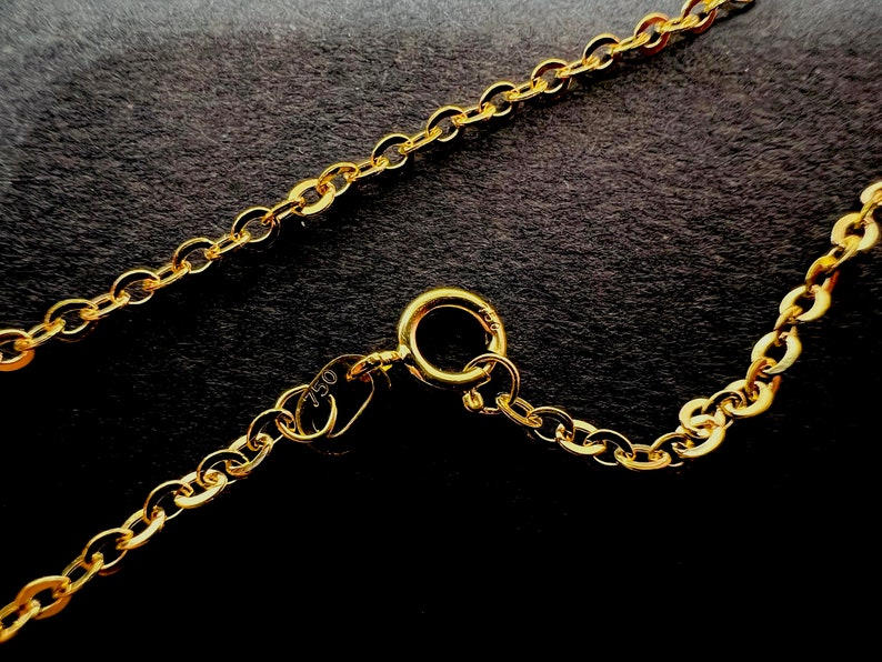 May include: Close-up of a gold necklace with an oval link chain. The necklace has a lobster clasp closure and a small tag with the number "750" stamped on it. The chain is displayed against a dark background.
