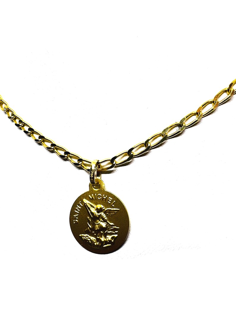 Puede incluir: Un collar dorado con un colgante que representa a San Miguel. El colgante redondo tiene las palabras "SAINT MICHEL" alrededor de la imagen. La cadena es una cadena figaro dorada.