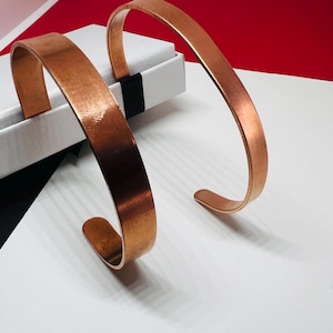 Peut inclure: Deux bracelets ouverts de type jonc fabriqués en cuivre poli avec une finition métallique lisse. Chaque bracelet présente un design simple en forme de bande incurvée avec des extrémités arrondies, mettant en valeur la teinte cuivrée.