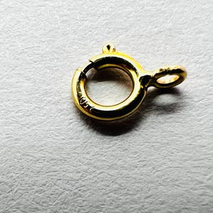 Può includere: Una chiusura a molla rotonda in oro lucido con un piccolo meccanismo a leva sporgente e un anellino di giunzione. La superficie metallica presenta un marchio impresso con la scritta 750, che indica la purezza dell'oro del componente.