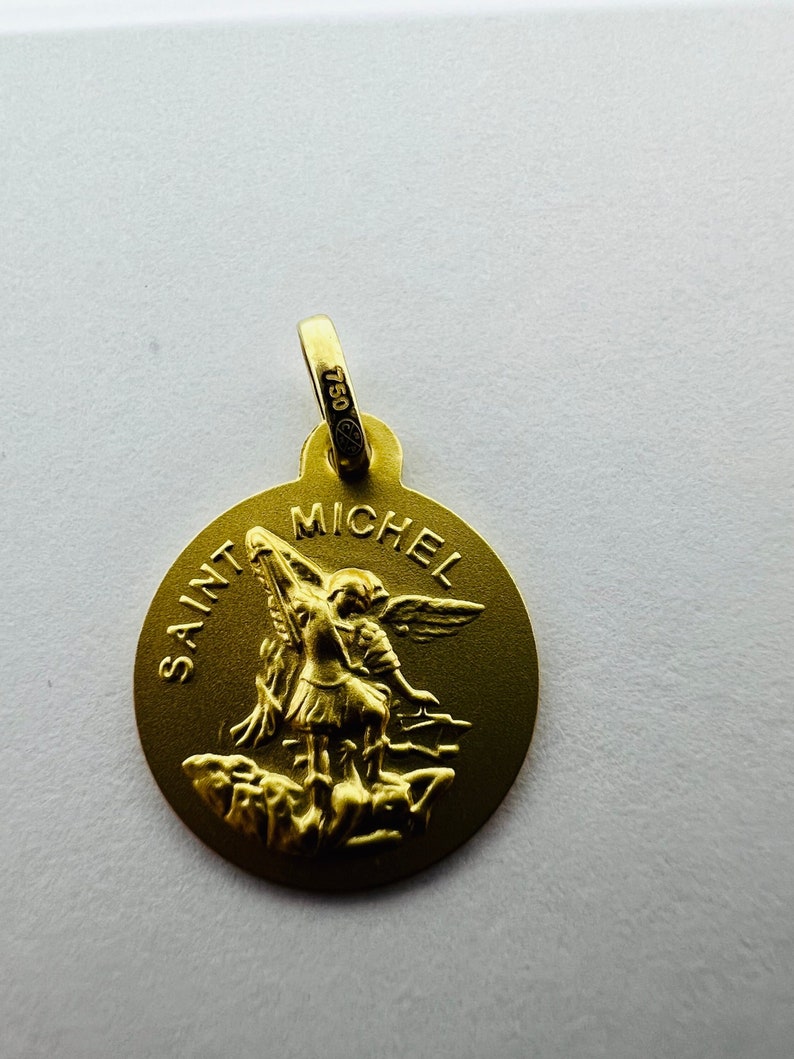 Puede incluir: Un colgante dorado con las palabras "SAINT MICHEL" y la imagen de un &aacute;ngel. El colgante tiene un lazo en la parte superior con el n&uacute;mero "750" estampado. El colgante es redondo y tiene un acabado mate.