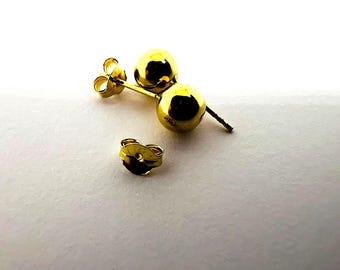Può includere: Un paio di orecchini a lobo dorati a sfera con chiusura a farfalla. Gli orecchini sono su una superficie bianca. Design semplice e classico.