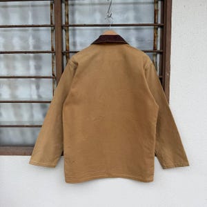 Puede incluir: Una chaqueta de trabajo marr&oacute;n con cuello de pana marr&oacute;n cuelga de una percha de madera. La chaqueta tiene mangas largas y una botonadura frontal. La parte trasera de la chaqueta es visible, mostrando las costuras y los detalles de construcci&oacute;n.