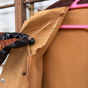Puede incluir: Primer plano de una chaqueta de trabajo color canela con cuello de pana marr&oacute;n. La chaqueta tiene cierre de bot&oacute;n y un aspecto desgastado y usado. El forro interior es visible y la chaqueta est&aacute; sujeta por una mano enguantada.
