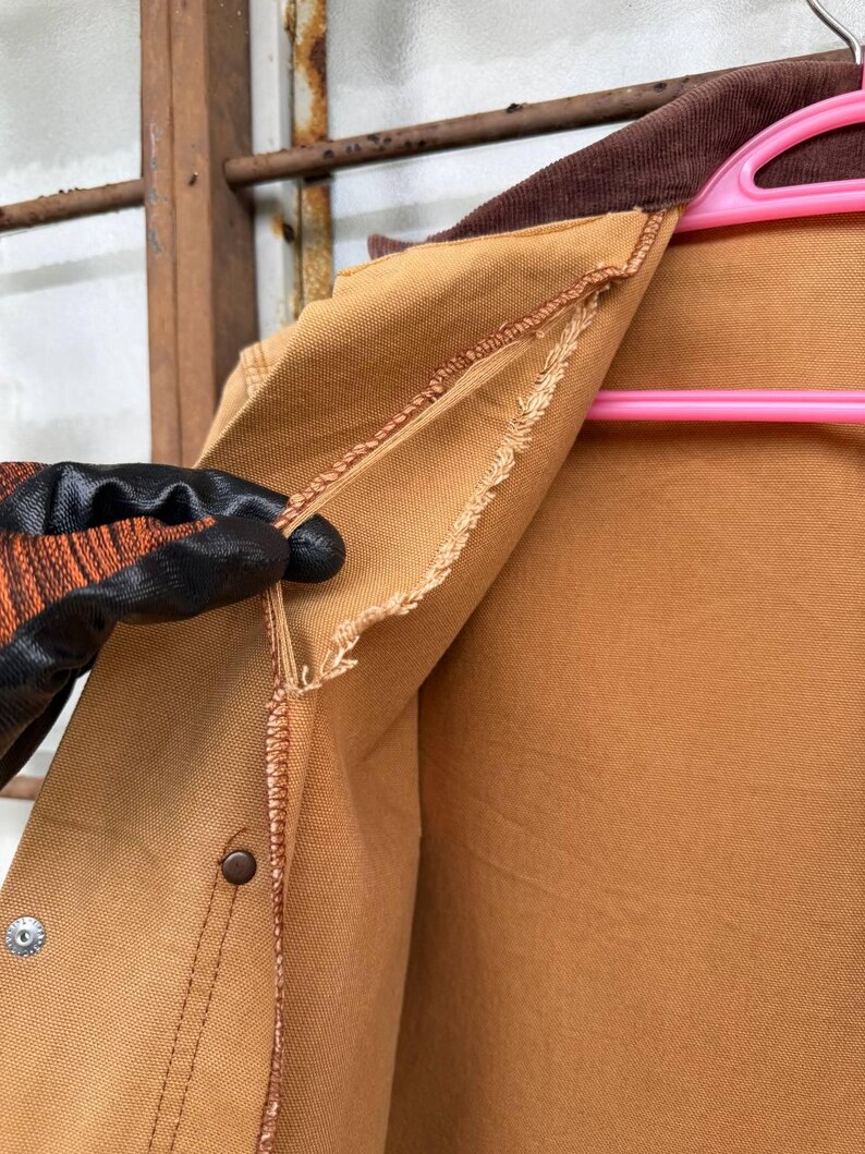 Puede incluir: Primer plano de una chaqueta de trabajo color canela con cuello de pana marr&oacute;n. La chaqueta tiene cierre de bot&oacute;n y un aspecto desgastado y usado. El forro interior es visible y la chaqueta est&aacute; sujeta por una mano enguantada.