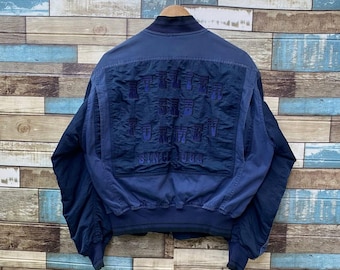 May include: A navy blue bomber jacket with a ribbed collar and cuffs. The back features a textured panel with purple text that reads "SINCE 1984". The jacket is hanging on a wooden hanger against a multi-colored wall.
