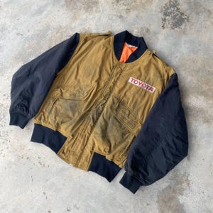 May include: A brown and black bomber jacket with a black zipper and a patch that says "TOYOTA" in red letters.