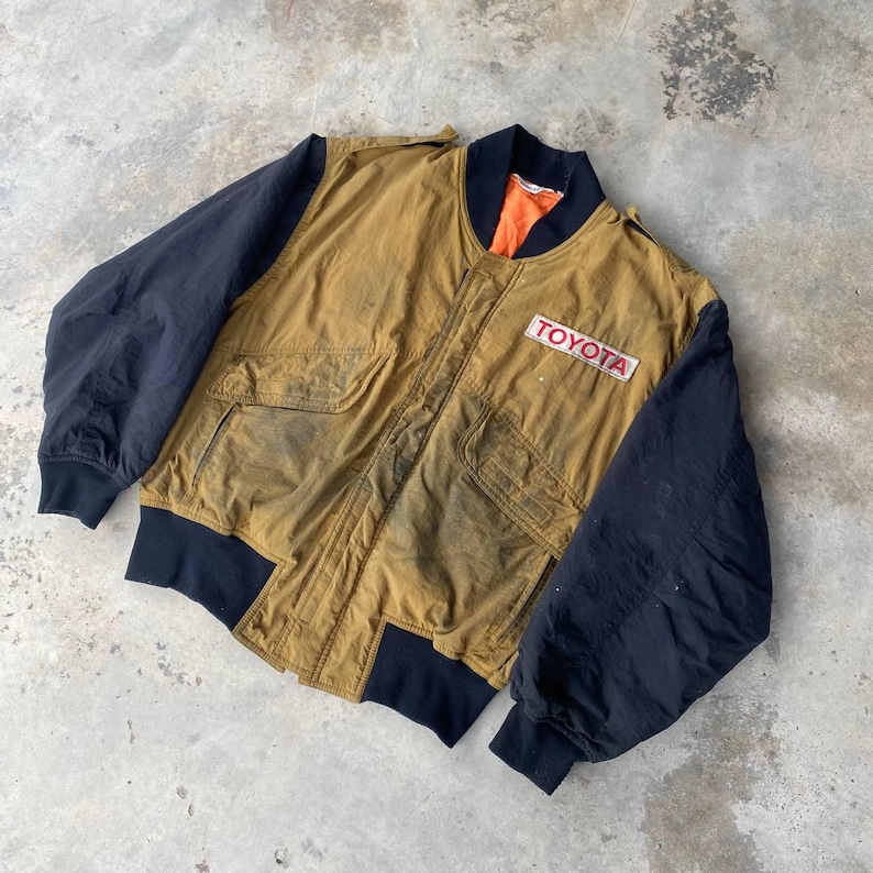 May include: A brown and black bomber jacket with a black zipper and a patch that says "TOYOTA" in red letters.