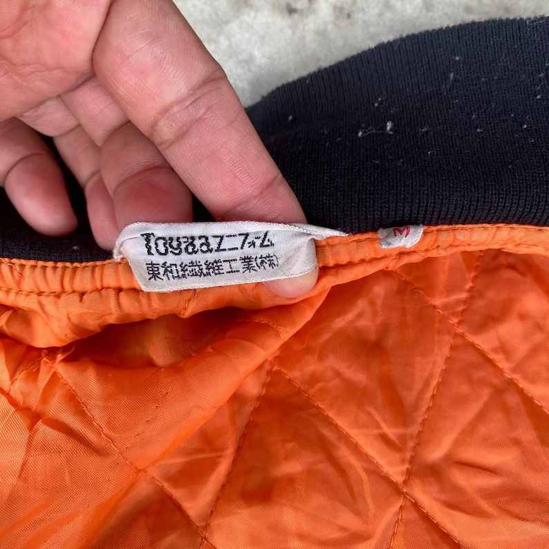 May include: Close-up of a black and orange bomber jacket with a white label that reads "Toy's Dream" and "東和縫製工業(株)".