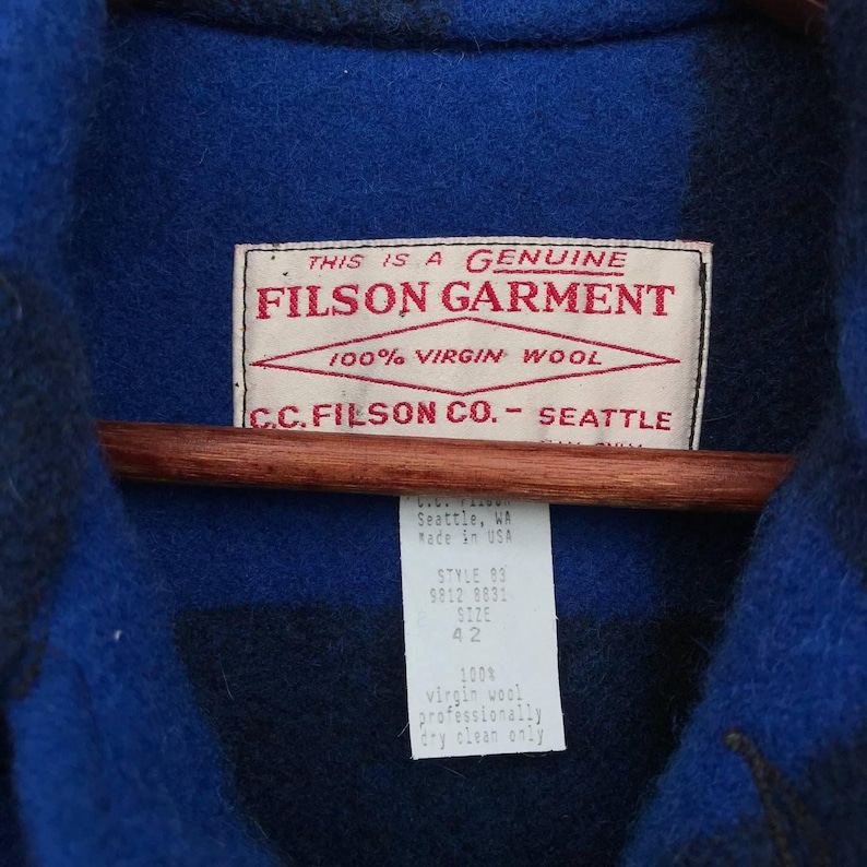 Puede incluir: Primer plano de una prenda Filson de lana azul y negra. La etiqueta dice "FILSON GARMENT" y "100% VIRGIN WOOL". Una etiqueta indica la talla 42 y que la prenda est&aacute; hecha en EE. UU. Limpieza en seco profesional solamente.