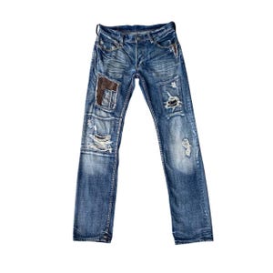 Peut inclure: Jean bleu en denim avec des détails usés, notamment des déchirures et des patchs. Le jean a une braguette boutonnée et une conception classique à cinq poches. Les patchs sont marron. Le jean est posé à plat sur fond blanc.
