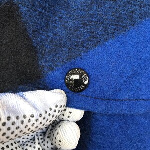 Puede incluir: Primer plano de una tela a cuadros azul y negro con un bot&oacute;n negro. El bot&oacute;n es redondo y tiene texto en relieve. La tela es texturizada y parece ser lana o un material similar.