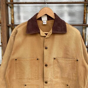 Puede incluir: Chaqueta de trabajo color canela con cuello de pana marr&oacute;n, cierre de botones y cuatro bolsillos delanteros. La chaqueta est&aacute; colgada de una percha de madera. Se ve una peque&ntilde;a etiqueta con la bandera americana.
