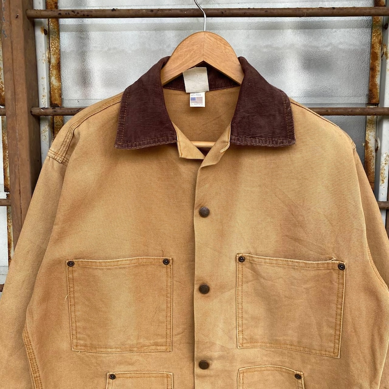 Puede incluir: Chaqueta de trabajo color canela con cuello de pana marr&oacute;n, cierre de botones y cuatro bolsillos delanteros. La chaqueta est&aacute; colgada de una percha de madera. Se ve una peque&ntilde;a etiqueta con la bandera americana.