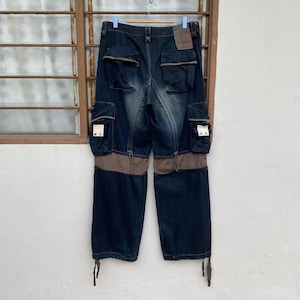 May include: Dark wash denim cargo pants with multiple pockets and brown accents. The pants have a relaxed fit and feature a unique design with contrasting fabric panels and drawstrings at the hem. A brown leather tag is visible.