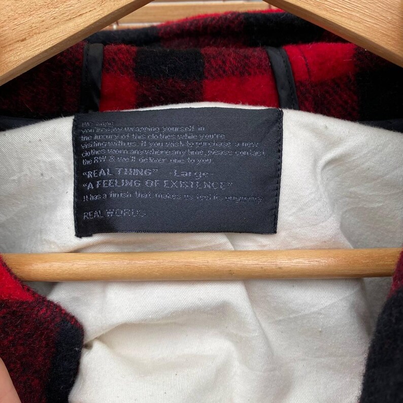 May include: A black and red plaid flannel shirt with a black tag that reads "REAL THING" "A FEELING OF EXISTENCE" REAL WORDS. The tag is attached to the shirt with a white thread.