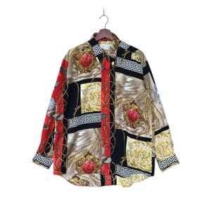 May include: A button-down shirt with a colorful pattern. The shirt features a mix of red, black, tan, and gold colors. The design includes ornate gold chains, red heart-shaped emblems, and Greek key borders. The long-sleeved shirt is hanging on a wooden hanger.