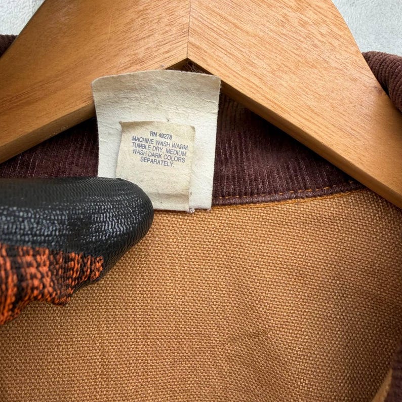 Puede incluir: Primer plano de una camisa de pana marr&oacute;n con una etiqueta que dice "RN 48278 MACHINE WASH WARM TUMBLE DRY MEDIUM WASH DARK COLORS SEPARATELY". La camisa est&aacute; colgada en una percha de madera.