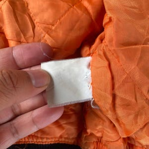 May include: Close-up of the orange lining of a jacket. The lining is quilted and has a white tag attached to it.