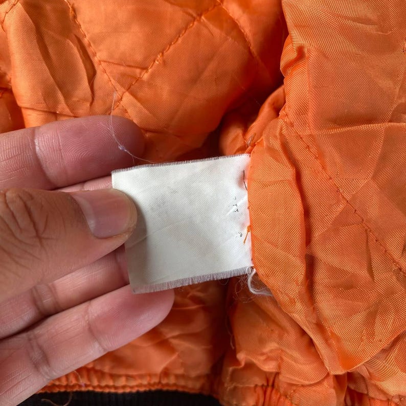 May include: Close-up of the orange lining of a jacket. The lining is quilted and has a white tag attached to it.
