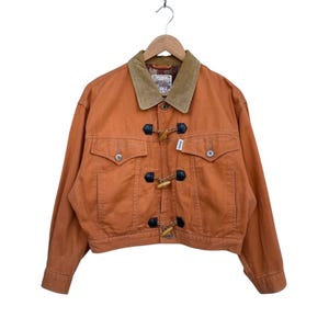 May include: An orange-colored jacket with a brown corduroy collar and toggle closures. The jacket has two front pockets and button details. The jacket is hanging from a wooden hanger.