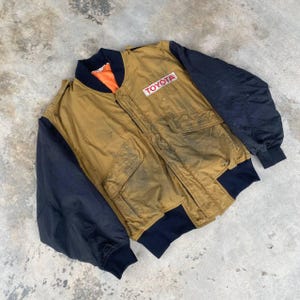 May include: A vintage brown and black bomber jacket with a large "TOYOTA" patch on the chest. The jacket has a zipper closure and two pockets.