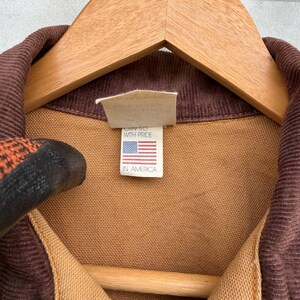 Puede incluir: Primer plano de un cuello de pana marr&oacute;n y tela beige de una chaqueta. Una etiqueta dice "Crafted with Pride" y presenta la bandera estadounidense y las palabras "In America". La chaqueta est&aacute; colgada en una percha de madera.